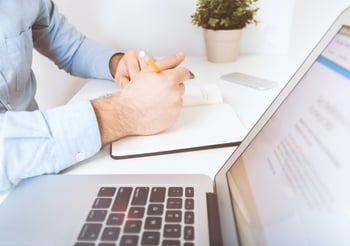 Person writing in a book to the left of an open laptop.