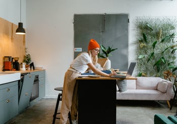 Woman wearing a red beanie standing over a countertop working on a laptop.
