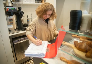 Woman in an industrial kitchen writing in a notebook.