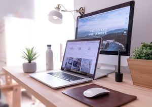 Desk setup with a laptop computer in front of a computer monitor, with a plant and water bottle to the left and a white mouse on a black mouse pad to the right.