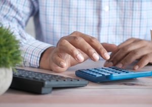 Person typing into a blue calculator next to a desktop keyboard.