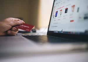 Person holding a red credit card in their left hand while shopping online on a laptop computer.