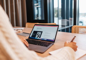 A person takes notes with a pen and notepad at a wooden table while working on a laptop computer.