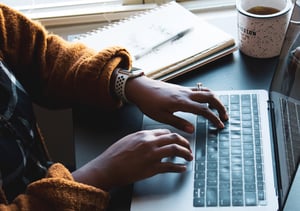 Person with an orange sweater typing on a laptop computer. next to a notebook and pen and a cup of coffee.Person with an orange sweater typing on a laptop computer. next to a notebook and pen and a cup of coffee.