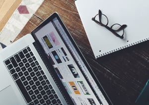 A silver laptop, notepad, and glasses on top of a wooden table.