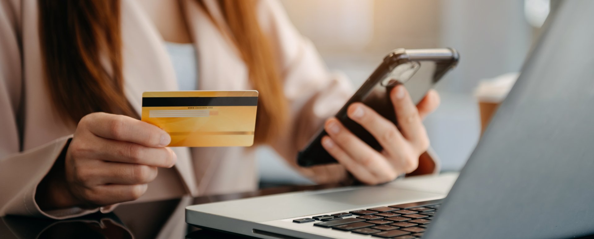 Person holding a smartphone in their left hand and a gold credit card in their right hand while sitting at a table looking at a laptop computer.