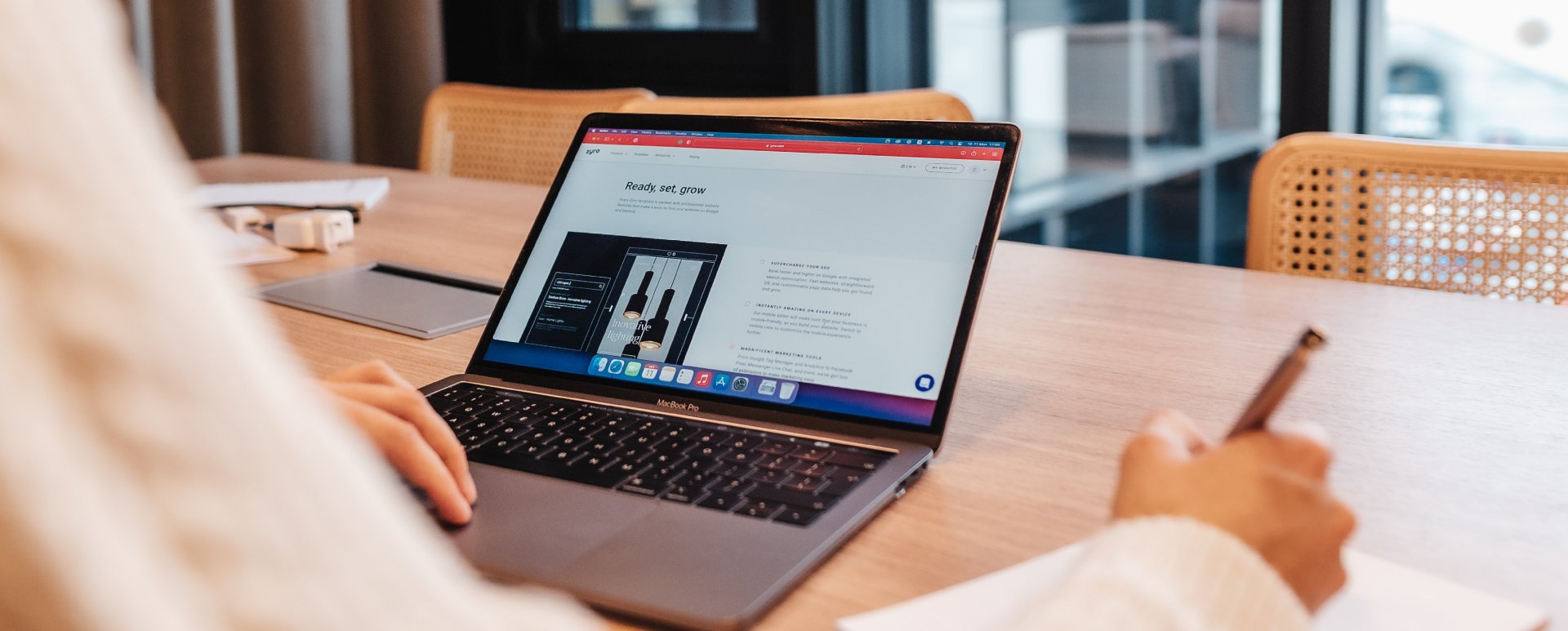 Person holding a pen and writing on a piece of paper while looking at a grey laptop.