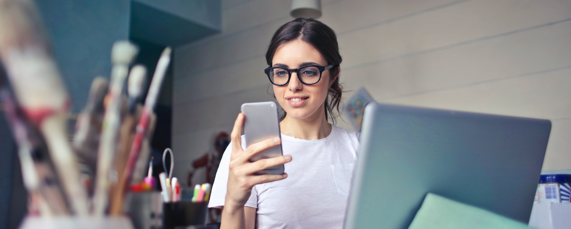 Woman wearing glasses looking at a cell phone while sitting in front of a computer.
