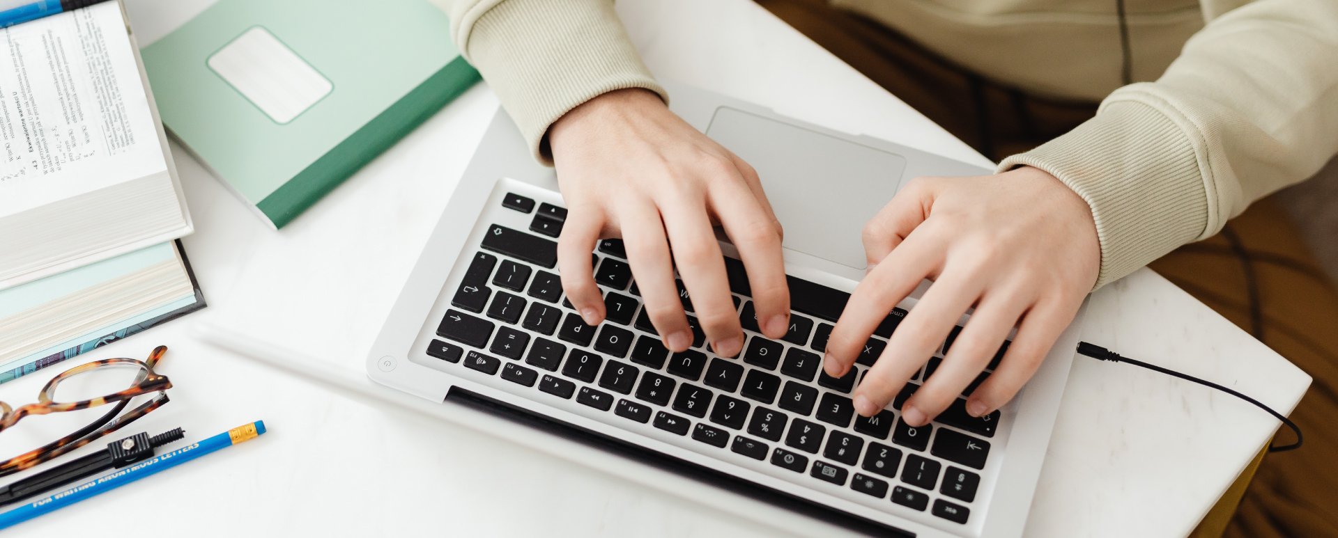 Person typing on a silver laptop computer on top of a white table.